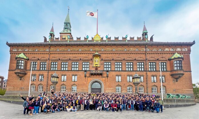 Una gran fotografía grupal que reúne a decenas de distribuidores, fabricantes y directivos de CT Internacional posando sonrientes y saludando frente a la imponente fachada histórica de ladrillo rojo del Ayuntamiento de Copenhague. En la parte superior del edificio, ondea una bandera blanca con el logotipo de la empresa organizadora bajo un cielo parcialmente nublado.