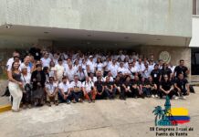 Congreso MyBusiness POS: XV años de un encuentro lleno de oportunidades Fotografía nocturna en plano general de un letrero monumental con letras tridimensionales iluminadas que forman la palabra "CARTAGENA" sobre una plataforma en la playa. Cada letra está decorada con pinturas artísticas y coloridas que muestran paisajes y rostros caribeños. En la esquina superior izquierda se superpone un logotipo cuadrangular blanco con una ilustración de una torre de reloj y una palma junto a la bandera de Colombia, detallando el texto "XV Congreso Anual de Punto de Venta - CARTAGENA | 28 AL 31 DE MAYO 2026 - MyBusiness POS".