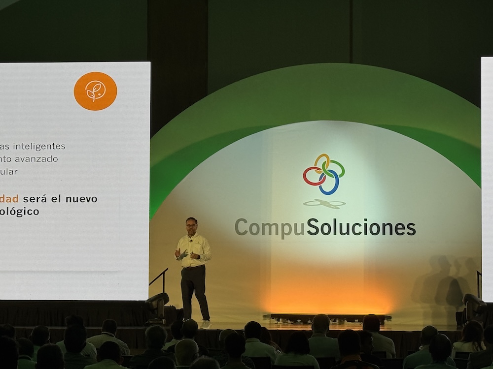 Encuadre medio de Juan Pablo Medina discursando en el escenario de un centro de convenciones repleto de asistentes sentados de espaldas. Se ubica a la izquierda de un monumental arco blanco que porta el imagotipo multicolor de CompuSoluciones, bajo una iluminación de acento en tonos amarillos y verdes.