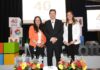 Microsoft 40 años en México impulsa la evolución tecnológica Rafael Sánchez posa de pie y sonriente en el centro del escenario, flanqueado por dos ejecutivas de la empresa. La mujer de la izquierda viste un saco naranja brillante y pantalón negro, mientras que la de la derecha porta un blazer blanco y pantalón oscuro. En primer plano, sobre una mesa, se muestra un pastel de aniversario decorado en blanco y azul junto a cuatro arreglos florales de rosas y gerberas, con la tipografía gigante de Microsoft de fondo.