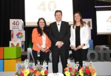 Microsoft 40 años en México impulsa la evolución tecnológica Rafael Sánchez posa de pie y sonriente en el centro del escenario, flanqueado por dos ejecutivas de la empresa. La mujer de la izquierda viste un saco naranja brillante y pantalón negro, mientras que la de la derecha porta un blazer blanco y pantalón oscuro. En primer plano, sobre una mesa, se muestra un pastel de aniversario decorado en blanco y azul junto a cuatro arreglos florales de rosas y gerberas, con la tipografía gigante de Microsoft de fondo.