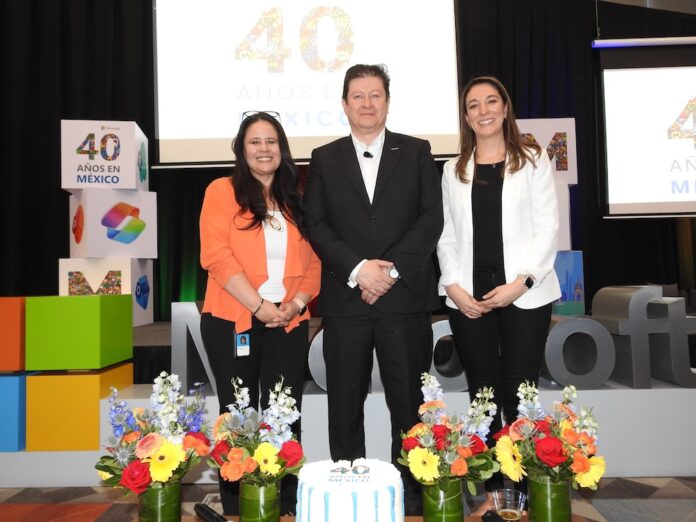 Rafael Sánchez posa de pie y sonriente en el centro del escenario, flanqueado por dos ejecutivas de la empresa. La mujer de la izquierda viste un saco naranja brillante y pantalón negro, mientras que la de la derecha porta un blazer blanco y pantalón oscuro. En primer plano, sobre una mesa, se muestra un pastel de aniversario decorado en blanco y azul junto a cuatro arreglos florales de rosas y gerberas, con la tipografía gigante de Microsoft de fondo.