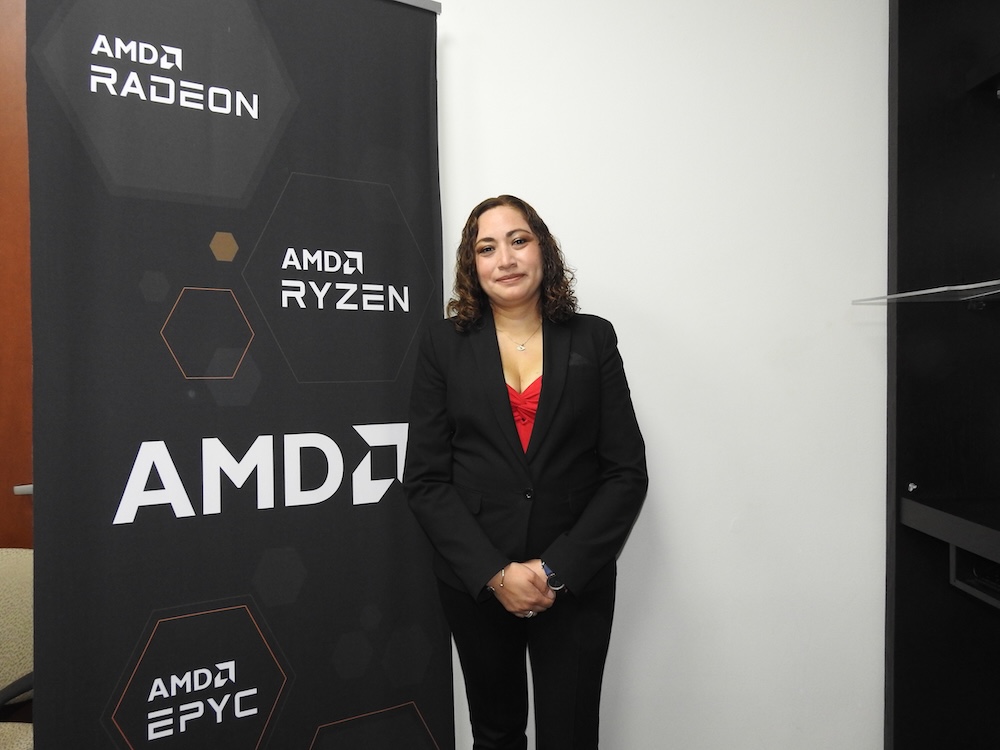 Retrato de Gabriela Alvarado, SMB Manager para Centroamérica y Caribe en AMD, de pie junto a un tótem publicitario con los logos de AMD Ryzen, Radeon y EPYC.