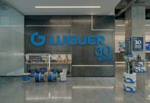 Grupo Luguer impulsa oportunidades enterprise para el canal TI Instalaciones del laboratorio de entrenamiento técnico y cableado estructurado en las oficinas de Grupo Luguer. En la pared del fondo, un gran panel metálico de color gris oscuro exhibe el logotipo de la compañía en tipografía azul junto al texto conmemorativo "30 años". Abajo, en los mostradores y en el suelo de concreto pulido, se disponen rollos de cable Categoría 6A de color azul, cajas de cartón con el emblema de la empresa y maniquíes de entrenamiento para la certificación técnica de integradores.