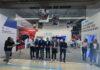 Centro de experiencia HP abre una nueva etapa para la experiencia tecnológica en retail Perspectiva general desde el pasillo comercial hacia la ceremonia de inauguración del Centro de Experiencia. En el centro, una fila de seis directivos de HP e Intel cortan de forma sincronizada una cinta roja delgada con tijeras plateadas, acompañados a la izquierda por dos fotógrafos que registran el evento y a la derecha por dos ejecutivas de relaciones públicas, una de ellas hablando por un micrófono. El stand blanco del fondo exhibe letreros con los títulos "Descubre el poder de la innovación" y "Centro de Experiencias" bajo los logotipos de ambas firmas.