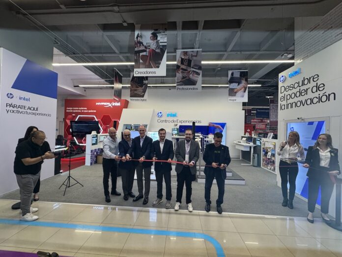HP e Intel realizan el corte de listón de su Centro de Experiencia interactivo Perspectiva general desde el pasillo comercial hacia la ceremonia de inauguración del Centro de Experiencia. En el centro, una fila de seis directivos de HP e Intel cortan de forma sincronizada una cinta roja delgada con tijeras plateadas, acompañados a la izquierda por dos fotógrafos que registran el evento y a la derecha por dos ejecutivas de relaciones públicas, una de ellas hablando por un micrófono. El stand blanco del fondo exhibe letreros con los títulos