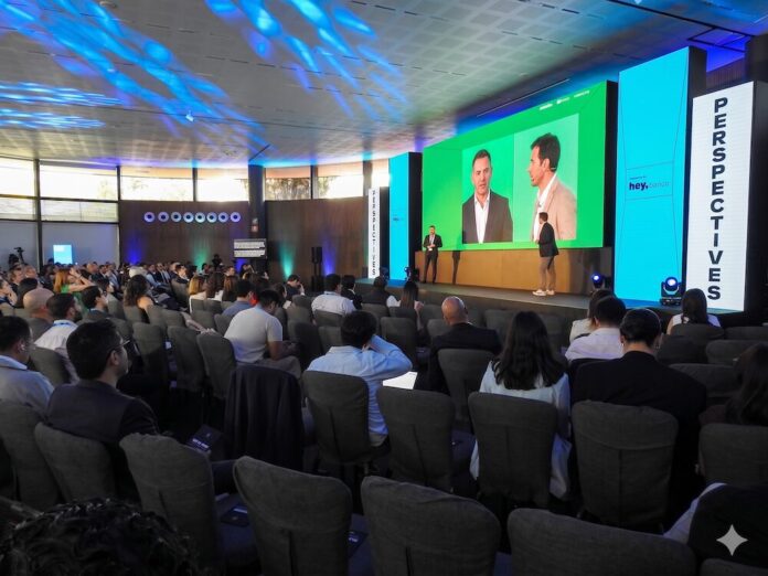 Hey Banco encabeza el encuentro empresarial Perspectives Auditorio lleno durante el foro Perspectives presentado por Hey Banco, mostrando a los asistentes desde atrás presenciando la conferencia de los ponentes en el escenario principal bajo una iluminación azul de techo.