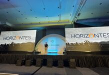CompuSoluciones Horizontes impulsa la IA en canales Perspectiva general desde el público del escenario principal de una convención empresarial iluminada con luces azules. En el centro, bajo una estructura de arco iluminada con el logotipo de CompuSoluciones, Juan Pablo Medina habla con un micrófono en la mano, flanqueado a los costados por dos monumentales pantallas LED panorámicas que proyectan la palabra "HORIZONTES" sobre un fondo de paisaje montañoso en blanco y negro.