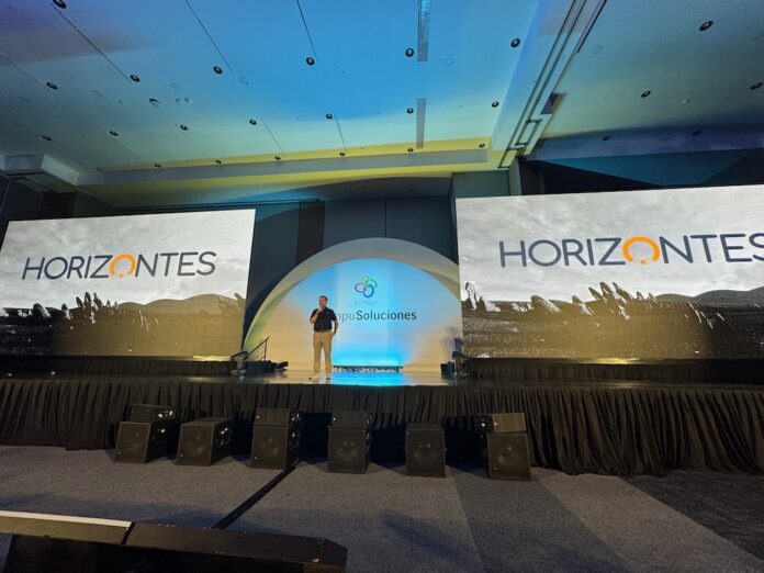 Perspectiva general desde el público del escenario principal de una convención empresarial iluminada con luces azules. En el centro, bajo una estructura de arco iluminada con el logotipo de CompuSoluciones, Juan Pablo Medina habla con un micrófono en la mano, flanqueado a los costados por dos monumentales pantallas LED panorámicas que proyectan la palabra 