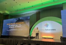CompuSoluciones traza el rumbo del canal con 8 macro-tendencias y la evolución del negocio de distribución Perspectiva general desde el público del escenario principal de un centro de convenciones iluminado con tonalidades verdes y naranjas. En el proscenio, bajo un arco blanco iluminado que exhibe el logotipo de CompuSoluciones, un orador discursa de pie con el micrófono en la mano, flanqueado a su izquierda por una pantalla monumental que despliega el texto "Horizontes de crecimiento: El modelo que nos proyecta al futuro - Juan Pablo Medina Mora".