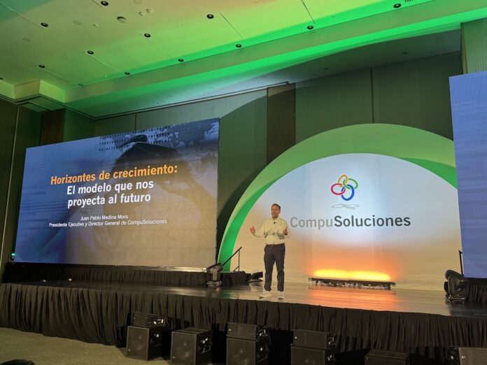 Juan Pablo Medina presenta las seis estrategias clave para transformar el canal de distribución TI Perspectiva general desde el público del escenario principal de un centro de convenciones iluminado con tonalidades verdes y naranjas. En el proscenio, bajo un arco blanco iluminado que exhibe el logotipo de CompuSoluciones, un orador discursa de pie con el micrófono en la mano, flanqueado a su izquierda por una pantalla monumental que despliega el texto