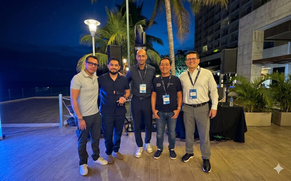 Fotografía grupal de plano entero tomada por la noche en un entorno tropical con palmeras de fondo. Cinco ejecutivos varones posan alineados y sonrientes de pie sobre un piso de madera clara. Visten ropa casual formal, destacando el segundo de izquierda a derecha con camisa negra, quien sostiene el trofeo de Netskope otorgado a Licencias OnLine. Tres de los directivos centrales portan gafetes celestes colgados al cuello y el ejecutivo de la extrema derecha usa lentes y camisa blanca con pantalón gris.