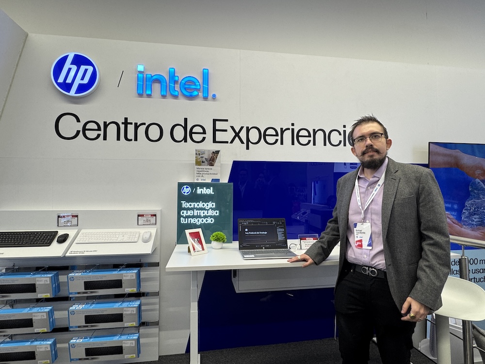 Retrato de plano medio y de frente de Rafael Escalante, Director de Tecnología de Intel, posando sonriente con la mano derecha apoyada sobre una mesa blanca. Viste un blazer formal gris oxford sobre una camisa clara de cuello abierto, lentes de armazón negro y porta un gafete colgante de identificación, situándose de pie por delante de una computadora portátil HP y debajo del letrero corporativo luminoso de "hp / intel Centro de Experiencia".