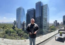 Licencias OnLine y Netskope fortalecen su alianza tras reconocimiento regional y participación estratégica Retrato de plano medio de frente del ingeniero Tomás Pérez, especialista técnico de Licencias OnLine, posando de pie en una terraza al aire libre bajo la luz del día. Sostiene con ambas manos un trofeo acrílico negro transparente con bordes redondeados que lleva impreso el logotipo de Netskope, seguido del texto "LICENCIAS ONLINE - Best technical partner FY2026". El ingeniero viste una camisa de mezclilla gris oscuro y pantalón de mezclilla claro, teniendo a sus espaldas una barandilla de cristal que revela un fondo urbano de rascacielos modernos de cristal azul bajo un cielo despejado.