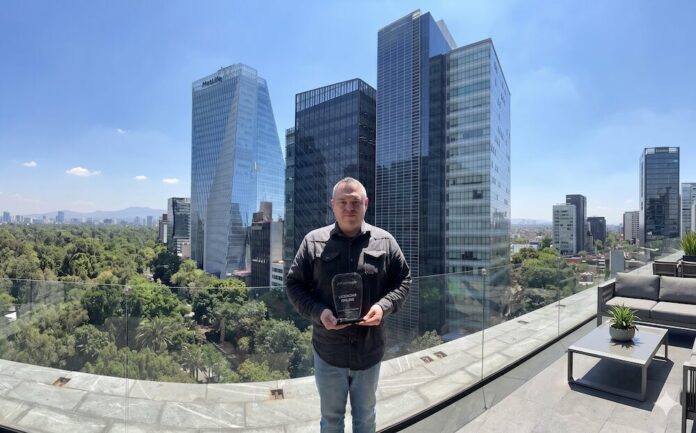 Tomás Pérez recibe el galardón al mejor socio técnico en el Partner Advisory Council de Netskope Retrato de plano medio de frente del ingeniero Tomás Pérez, especialista técnico de Licencias OnLine, posando de pie en una terraza al aire libre bajo la luz del día. Sostiene con ambas manos un trofeo acrílico negro transparente con bordes redondeados que lleva impreso el logotipo de Netskope, seguido del texto