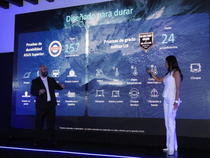 Fotografía de plano general en un escenario iluminado donde Francisco Jiménez y Paola Rojas presentan ante una gran pantalla de proyección digital. Francisco viste un traje oscuro y camisa blanca, apuntando con la mano hacia las gráficas, mientras Paola, con un conjunto de blusa y pantalón completamente blancos, sostiene un micrófono. La pantalla despliega el título Diseñado para durar junto con íconos informativos que detallan pruebas de durabilidad ASUS Superior y de grado militar US MIL-STD 810H.
