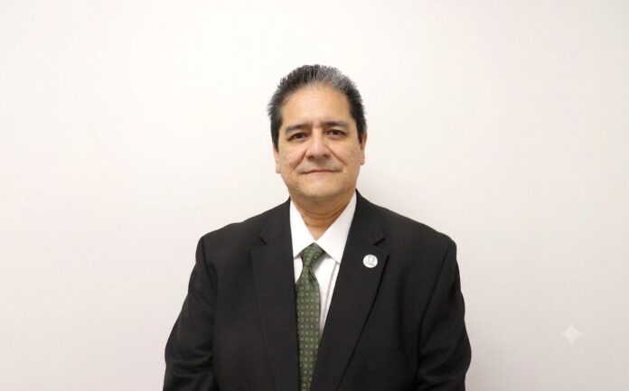 Fotografía de estudio en plano medio que muestra a Alberto Escamilla, Director General de Fulfillment Management México (FMM), posando de frente sobre un fondo completamente blanco. El directivo tiene tez clara y cabello corto oscuro con sutiles canas en las sienes, y viste un saco formal negro entallado sobre una camisa de vestir blanca y una corbata verde oliva con patrones geométricos sutiles. En la solapa izquierda de su saco resalta un pequeño pin metálico con el logotipo circular de la empresa, manteniendo los brazos sueltos a los costados y una mirada sutil frente a la cámara.