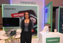 Kaspersky impulsa una nueva visión de la ciberseguridad para el hogar Fotografía en plano medio de Angélica Ruiz, Account Manager para B2C en Kaspersky, posando de frente y con las manos en los bolsillos dentro de un stand de exposición. La ejecutiva utiliza anteojos de armazón oscuro, un collar con un dije circular y viste un chaleco sastre negro combinado con una blusa gris de manga larga y pantalones negros. A su izquierda se observa un mostrador blanco con el logotipo de la marca, mientras que a su espalda destaca una pantalla publicitaria con la ilustración de un celular bloqueando spam y una mesa de cristal con computadoras portátiles abiertas.