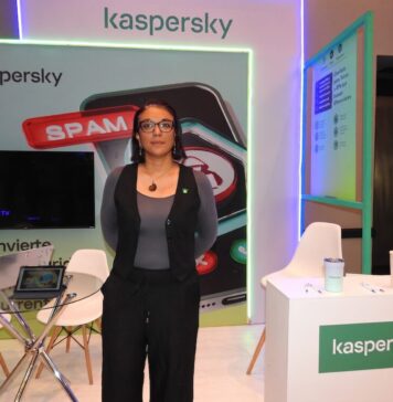 Kaspersky impulsa una nueva visión de la ciberseguridad para el hogar Fotografía en plano medio de Angélica Ruiz, Account Manager para B2C en Kaspersky, posando de frente y con las manos en los bolsillos dentro de un stand de exposición. La ejecutiva utiliza anteojos de armazón oscuro, un collar con un dije circular y viste un chaleco sastre negro combinado con una blusa gris de manga larga y pantalones negros. A su izquierda se observa un mostrador blanco con el logotipo de la marca, mientras que a su espalda destaca una pantalla publicitaria con la ilustración de un celular bloqueando spam y una mesa de cristal con computadoras portátiles abiertas.
