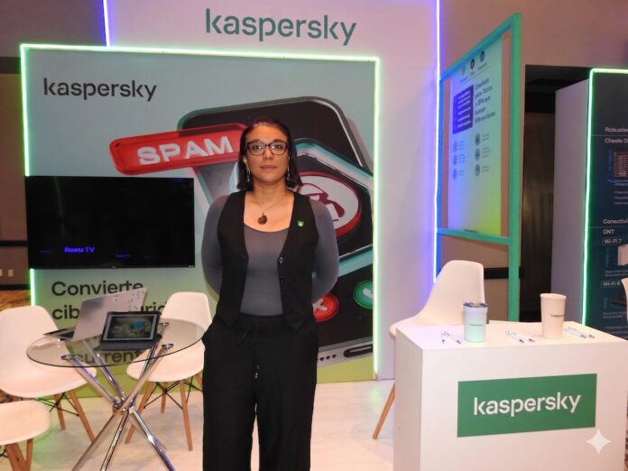 Fotografía en plano medio de Angélica Ruiz, Account Manager para B2C en Kaspersky, posando de frente y con las manos en los bolsillos dentro de un stand de exposición. La ejecutiva utiliza anteojos de armazón oscuro, un collar con un dije circular y viste un chaleco sastre negro combinado con una blusa gris de manga larga y pantalones negros. A su izquierda se observa un mostrador blanco con el logotipo de la marca, mientras que a su espalda destaca una pantalla publicitaria con la ilustración de un celular bloqueando spam y una mesa de cristal con computadoras portátiles abiertas.