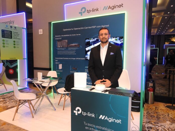 Benjamín del Río presenta el portafolio tecnológico de TP-Link y Aginet diseñado para optimizar operaciones Carrier e ISP en México Fotografía en plano medio largo que muestra a Benjamín del Río, Business Development Manager de TP-Link, posando de pie y sonriente en el stand de la marca durante la exposición Conecta. El ejecutivo tiene barba corta recortada y viste un traje sastre negro formal sobre una camisa blanca de cuello abierto con un pañuelo azul en el bolsillo superior izquierdo. Se ubica detrás de un mostrador promocional azul turquesa con el letrero