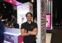 Bull Rocket: el auge de los agentes de IA para empresas transforma la automatización empresarial Fotografía en plano medio que muestra a Manuel Fernando Cobarrubias, coordinador de marketing de Bull Rocket, posando de frente con los brazos cruzados en el stand corporativo de la empresa durante el evento América Digital 2026. El profesional, de cabello oscuro rizado y anteojos de armazón transparente, viste una playera tipo polo negra con el logotipo bordado de "Bull Rocket". A la izquierda, al fondo, se observa a un compañero operando una computadora portátil sobre un mostrador blanco iluminado con luz LED rosa, mientras que a la derecha resalta una columna decorativa con textos publicitarios en blanco sobre fondo gris, junto a una pantalla vertical brillante que muestra la ilustración de un robot asistente de inteligencia artificial.