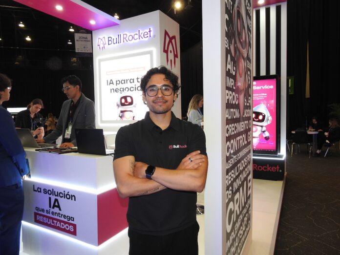 Fotografía en plano medio que muestra a Manuel Fernando Cobarrubias, coordinador de marketing de Bull Rocket, posando de frente con los brazos cruzados en el stand corporativo de la empresa durante el evento América Digital 2026. El profesional, de cabello oscuro rizado y anteojos de armazón transparente, viste una playera tipo polo negra con el logotipo bordado de 