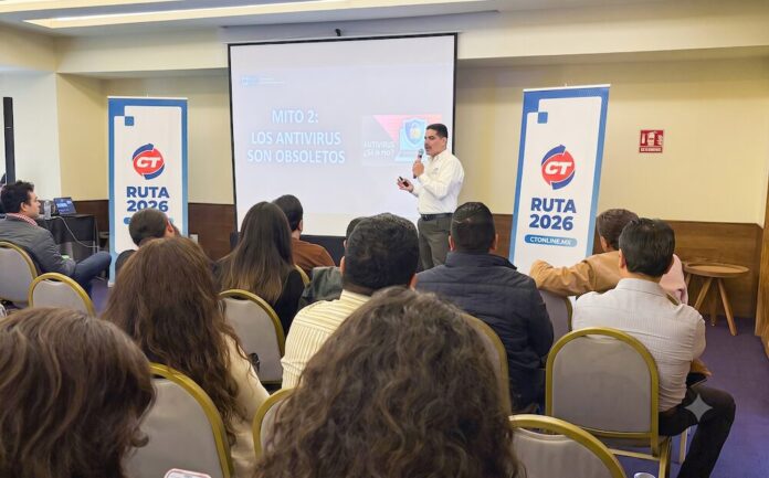Fotografía que muestra una sala de conferencias con un conferencista de pie hablando frente a una audiencia sentada de espaldas. El ponente, un hombre de camisa blanca y pantalón oscuro, sostiene un micrófono inalámbrico. A los lados de la pantalla de proyección se ubican dos tótems publicitarios azules con el logotipo de CT Internacional y la leyenda 