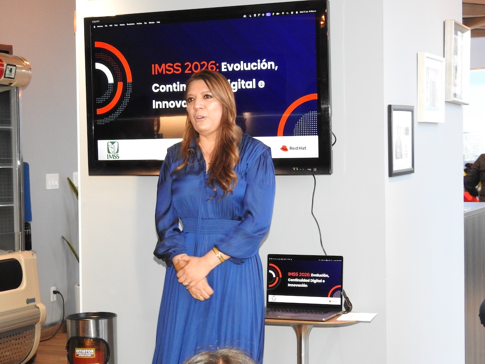 Fotografía en plano medio de Claudia Vázquez, Directora de Innovación del IMSS, de pie en una sala corporativa. Viste un vestido largo plisado de color azul rey y mantiene las manos entrelazadas al frente. Detrás de ella se ubica una pantalla de televisión montada en la pared que despliega la diapositiva "IMSS 2026: Evolución, Continuidad Digital e Innovación" con los logotipos de IMSS y Red Hat, replicada abajo en una computadora portátil abierta sobre una mesa circular.