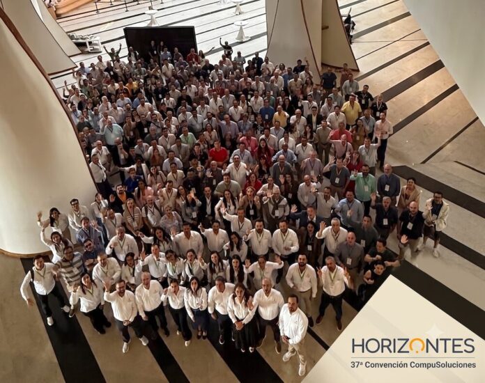 CompuSoluciones Horizontes reúne a su ecosistema de asociados en su convención anual Fotografía aérea en picada de un grupo masivo de más de cien asociados y profesionales congregados en el amplio vestíbulo de un centro de convenciones con pisos de mármol y grandes columnas blancas integradas. La mayoría de los asistentes mira hacia la cámara y saluda con la mano levantada. En el extremo inferior derecho se sobrepone un logotipo triangular de fondo blanco con la leyenda