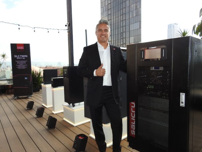 Consolidación de la alianza tecnológica entre Stulz y Salicru para el mercado de protección eléctrica en México Fotografía de frente en plano general medio de Emigdio Granillo, Managing Director de Stulz, posando en la terraza del evento al lado de una torre de energía negra de la marca Salicru. El directivo sonríe formalmente a la cámara y hace una señal de aprobación levantando el pulgar de su mano derecha. Viste un traje de vestir negro y una camisa blanca, estando rodeado en el fondo por pedestales con otros modelos de UPS más pequeños e indicadores publicitarios bajo un cielo claro de día.