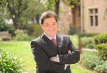 DXC fortalece su estrategia de alianzas en América Latina con el regreso de Emerson Dipardo Fotografía en plano medio que muestra a Emerson Dipardo posando de frente con una sonrisa y los brazos cruzados en un entorno exterior iluminado por luz natural. El ejecutivo viste un saco de traje negro, camisa de color azul claro, una corbata roja con rayas diagonales blancas y un reloj de pulsera con extensible metálico. El fondo se encuentra difuminado y revela un jardín con césped verde, vegetación arbórea, una banca de madera a la izquierda y parte de la fachada de un edificio de arquitectura clásica con ventanas de piedra a la derecha.