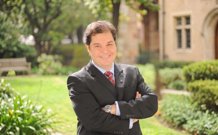 Fotografía en plano medio que muestra a Emerson Dipardo posando de frente con una sonrisa y los brazos cruzados en un entorno exterior iluminado por luz natural. El ejecutivo viste un saco de traje negro, camisa de color azul claro, una corbata roja con rayas diagonales blancas y un reloj de pulsera con extensible metálico. El fondo se encuentra difuminado y revela un jardín con césped verde, vegetación arbórea, una banca de madera a la izquierda y parte de la fachada de un edificio de arquitectura clásica con ventanas de piedra a la derecha.