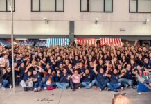 Los 20 años de Tecnosinergia: evolución y futuro de la seguridad electrónica Fotografía panorámica en plano general que muestra a todo el equipo de colaboradores de Tecnosinergia posando juntos para una foto grupal masiva en el patio exterior de sus instalaciones. Decenas de empleados aparecen sentados en el suelo y de pie con los brazos levantados en señal de festejo. El espacio cuenta con puestos o stands de feria con toldos de rayas rojas, blancas y azules, y en la esquina inferior derecha resalta un futbolito decorado con escudos deportivos.