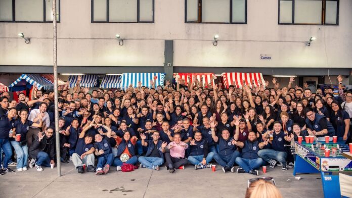 Fotografía panorámica en plano general que muestra a todo el equipo de colaboradores de Tecnosinergia posando juntos para una foto grupal masiva en el patio exterior de sus instalaciones. Decenas de empleados aparecen sentados en el suelo y de pie con los brazos levantados en señal de festejo. El espacio cuenta con puestos o stands de feria con toldos de rayas rojas, blancas y azules, y en la esquina inferior derecha resalta un futbolito decorado con escudos deportivos.