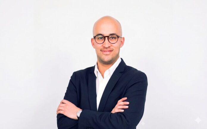 El VP of Growth de Auronix, analiza el impacto de las soluciones conversacionales transaccionales en la transformación empresarial Retrato de estudio en plano medio y de frente de un ejecutivo de complexión media, calvo y con lentes de armazón redondo oscuro, posando con una sonrisa ligera sobre un fondo gris claro uniforme. Viste un blazer negro formal bien entallado sobre una camisa blanca de vestir con cuello abierto. Mantiene los brazos cruzados al frente, dejando a la vista un reloj de pulsera plateado con correa negra en su muñeca izquierda.
