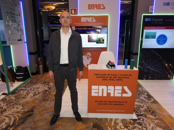 Fotografía de plano entero que muestra a Ricardo Ezeta, director operativo de Plásticos Enres, posando de pie junto al módulo comercial de su empresa en el congreso América Digital 2026 en la Ciudad de México. El directivo, calvo y con lentes, viste una camisa blanca de cuello abierto, una chamarra casual gris oscuro, pantalón oscuro y zapatos negros. A su izquierda destaca un mostrador rectangular de color naranja con el logotipo de 