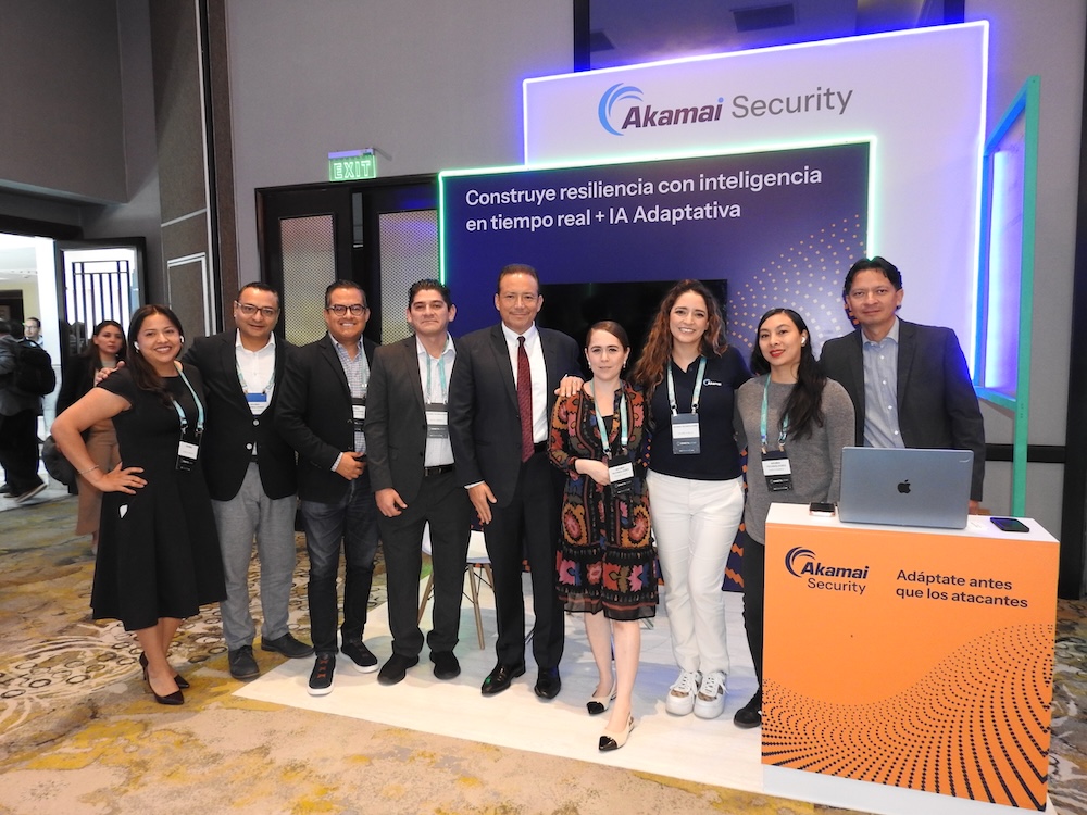 Servicios de ciberseguridad: Fotografía grupal en plano medio largo que muestra a nueve profesionales alineados de pie en el pabellón de Akamai Security dentro de una convención corporativa. Al centro destaca un ejecutivo de traje oscuro, camisa blanca y corbata roja, rodeado por cinco hombres y tres mujeres que portan credenciales del evento sobre vestimenta formal o casual de negocios. El stand de fondo cuenta con un letrero luminoso que dice "Construye resiliencia con inteligencia en tiempo real + IA Adaptativa", y en primer plano a la derecha se ubica un mostrador naranja con una computadora portátil cerrada.
