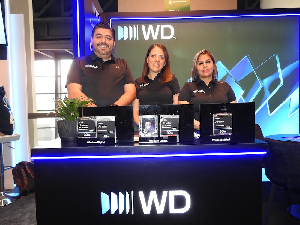 Almacenamiento empresarial interno seguro y escalable: Fotografía en plano medio que muestra a tres profesionales (un hombre a la izquierda y dos mujeres a la derecha) sonriendo detrás de un mostrador negro iluminado con tiras de luces LED azules y el logotipo tridimensional de WD. Los tres visten camisas polo negras con el emblema corporativo. Sobre la mesa se exhiben tres discos duros físicos de la línea Ultrastar de Western Digital, destacando capacidades de 30TB y 32TB sobre bases acrílicas oscuras. Al fondo, resalta un panel publicitario luminoso con diseños geométricos en tonos cian y azul.
