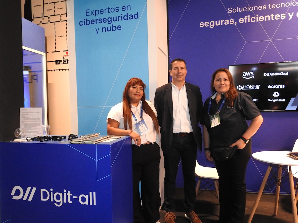 Ciberseguridad para empresas: Fotografía en plano general medio que muestra a tres colaboradores de la empresa Digit-all posando de frente en su stand corporativo. A la izquierda, una mujer de cabello largo castaño y blusa blanca se apoya en un mostrador azul brillante con el logotipo de "Digit-all". Al centro, un ejecutivo de saco oscuro y camisa blanca sonríe formalmente. A la derecha, otra colaboradora con playera polo negra complementa el grupo. El fondo del stand es azul rey y muestra un muro con la frase "Soluciones tecnológicas seguras, eficientes y..." junto a una pantalla digital que despliega logotipos de aliados como AWS, Alibaba Cloud, Honne Services, Acronis y Google Cloud.