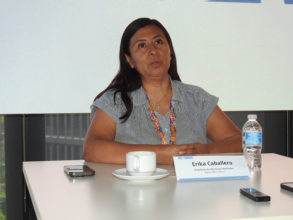 Fotografía en plano medio de Erika Caballero, Arquitecta de Soluciones DataCenter en Ingram Micro México, hablando durante una sesión informativa. Tiene el cabello largo oscuro y viste una blusa de rayas grises de manga corta con un cordón de acreditación multicolor. Está sentada detrás de una mesa blanca de oficina que contiene un teléfono celular, un identificador acrílico con su nombre y cargo, un bolígrafo y una botella de agua purificada al costado derecho, frente a una pantalla de proyección clara al fondo.