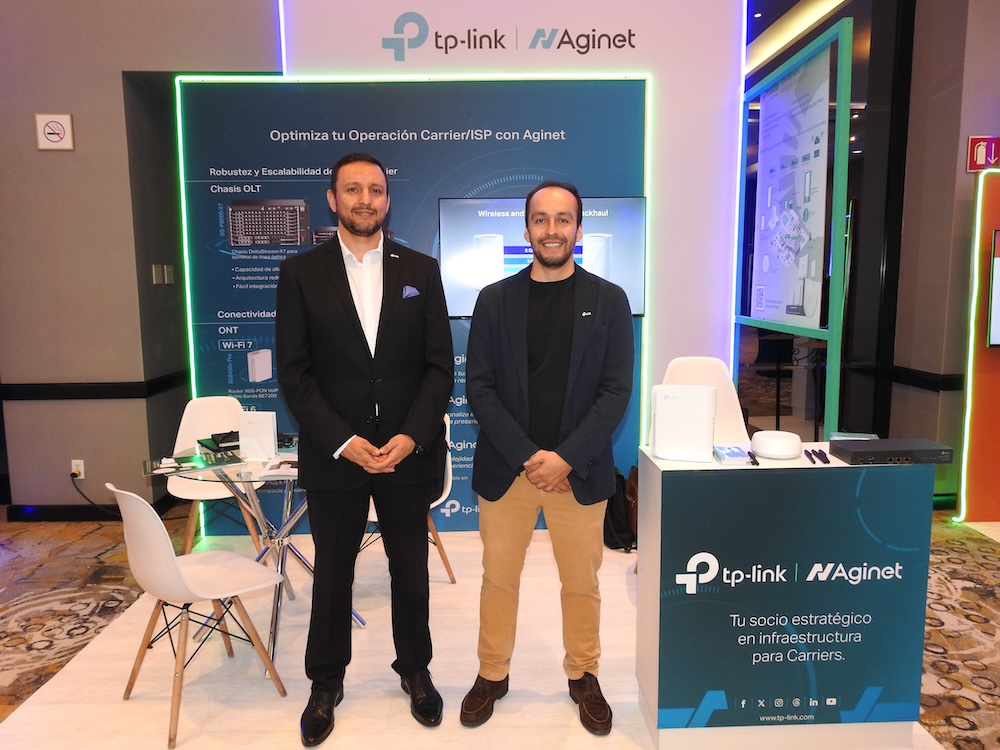 Conectividad ISP México: Fotografía en plano entero que muestra a dos ejecutivos de TP-Link posando juntos de pie y de frente dentro de un stand de exhibición tecnológica. El profesional de la izquierda es Benjamín del Río, quien viste un traje negro formal con camisa blanca. El colaborador de la derecha utiliza un saco sport azul marino sobre una playera negra, pantalones casuales de color beige claro y zapatos cafés. Ambos sonríen a la cámara flanqueados a la derecha por un mostrador informativo de color turquesa que muestra el texto en letras blancas "Tu socio estratégico en infraestructura para Carriers". Al fondo se observa el panel principal con iluminación LED de neón verde y el logotipo unificado de "tp-link | Aginet".