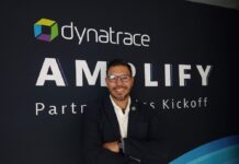 Dynatrace impulsa la observabilidad inteligente para las operaciones autónomas Fotografía en plano medio que muestra a Miguel Ángel Alarcón posando de frente con una sonrisa y los brazos cruzados ante un muro publicitario oscuro. El ejecutivo usa anteojos de armazón oscuro, una camisa blanca de cuello abierto y un saco sastre azul marino que lleva un pequeño pin multicolor con el logotipo de la marca en la solapa izquierda. El fondo negro muestra de manera prominente la palabra "AMPLIFY" debajo del logotipo de Dynatrace, seguido parcialmente por el texto en letras blancas "Partner Sales Kickoff".