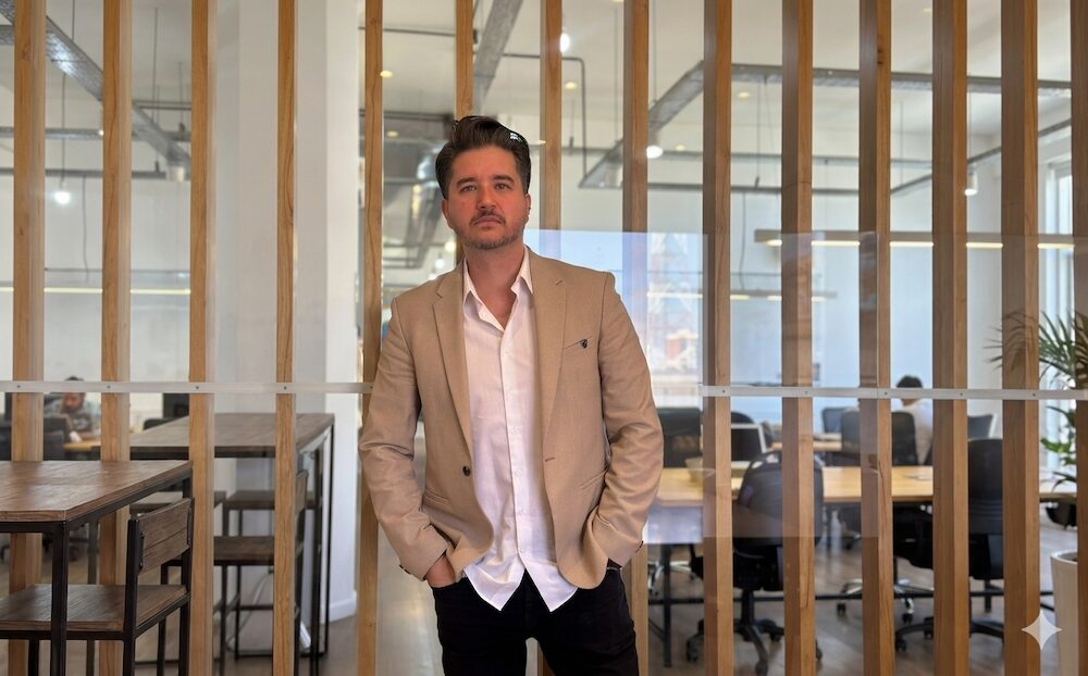 Retrato corporativo en plano medio de Fabián Perfetti, Co-Founder y Director de Streambe, posando con las manos en los bolsillos de su pantalón. Viste un saco de color beige claro sobre una camisa blanca de cuello abierto y pantalón negro. Se encuentra de pie en el interior de una oficina moderna, teniendo a sus espaldas un panel divisorio de barras verticales de madera clara y cristales que revelan un área de trabajo abierta con escritorios, computadoras y sillas ejecutivas.