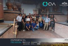 Grupo CVA reúne a fabricantes y distribuidores en Monterrey para fortalecer alianzas estratégicas Fotografía de plano entero que muestra a un grupo de aproximadamente quince personas posando de pie en el interior de una antigua planta de destilación artesanal con paredes de ladrillo rústico. Los asistentes, portando sombreros de paja, se ubican entre monumentales alambiques y estructuras cilíndricas de cobre brillante. En la parte inferior, se visualiza el banner promocional oscuro con el título "EXPERIENCIA PREMIUM" al lado de marcas como Microsoft Office y la firma de Casa Madero.