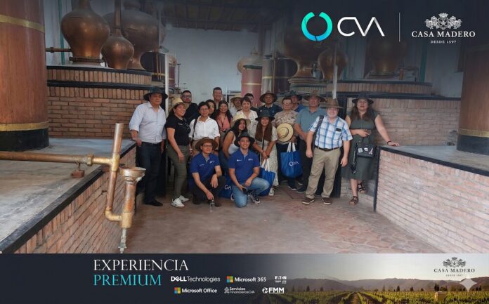 Fotografía de plano entero que muestra a un grupo de aproximadamente quince personas posando de pie en el interior de una antigua planta de destilación artesanal con paredes de ladrillo rústico. Los asistentes, portando sombreros de paja, se ubican entre monumentales alambiques y estructuras cilíndricas de cobre brillante. En la parte inferior, se visualiza el banner promocional oscuro con el título 
