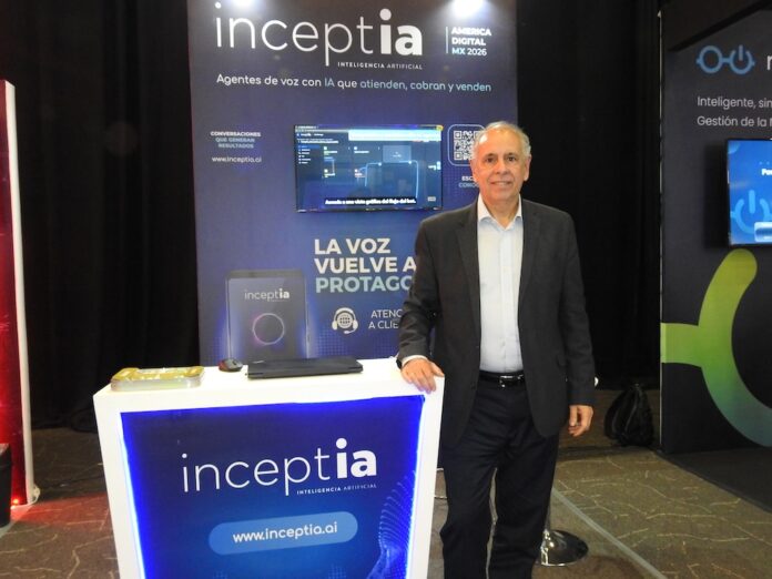 Fotografía en plano general medio de Gustavo Capart, CEO de Inceptia, posando de frente y sonriente junto al stand corporativo de su empresa durante el evento América Digital MX 2026. El directivo tiene cabello corto canoso, tez clara y viste un traje formal con saco gris oscuro sobre una camisa de vestir azul claro de cuello abierto, sin corbata, y pantalón oscuro. Apoya su mano derecha sobre un mostrador blanco retroiluminado con luces LED de color azul brillante que muestra el logotipo de 