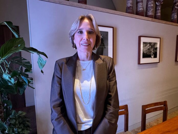 Retrato corporativo en plano medio y de frente de Adriana Flores posando en un entorno iluminado de forma tenue con tonalidades violetas. Viste un saco formal de color marrón satinado sobre una blusa blanca y luce collares de cadena delgada junto con aretes colgantes discretos. Mantiene una expresión sonriente y amable directa hacia la cámara, teniendo a sus espaldas una pared lisa decorada con cuadros fotográficos enmarcados y una planta de hojas grandes a la izquierda.