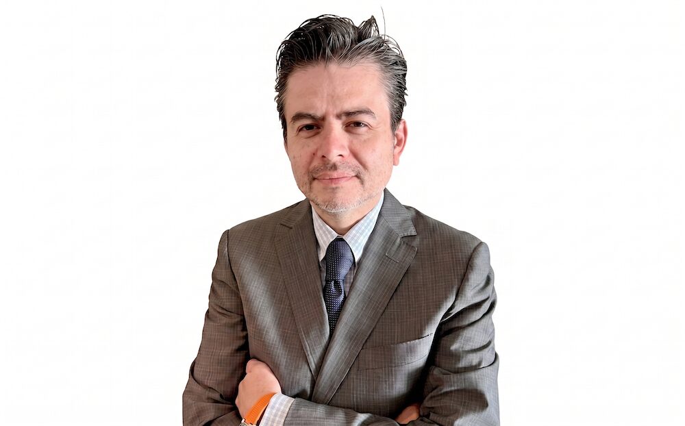 Retrato corporativo en plano medio de Herman Cipres, Business Development Manager (BDM) de Lenovo Global Financial Services, posando de frente con los brazos cruzados. Viste un saco formal gris con un sutil patrón de líneas sobre una camisa a cuadros claros y corbata oscura, portando un reloj con correa naranja en la muñeca izquierda. El sujeto se encuentra recortado de manera limpia sobre un fondo blanco totalmente liso y neutro.