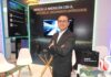 AMD lleva la Infraestructura de IA más allá de los centros de datos Fotografía en plano medio que muestra a Hiram Monroy, Sr Manager de AMD, posando de frente con los brazos cruzados en un stand corporativo. El ejecutivo viste un traje de color gris Oxford, camisa blanca y anteojos de armazón oscuro. Detrás de él, un muro publicitario negro con bordes iluminados por luces LED verdes exhibe la inscripción "IMPULSA LA INNOVACIÓN CON IA, ACELERA EL CRECIMIENTO EMPRESARIAL", junto a una pantalla digital central. A los lados se aprecian sillas de diseño blancas, una mesa de cristal y artículos promocionales de la marca que incluyen una bolsa negra con el lema "together we advance_AI", una playera de fútbol y un balón deportivo.